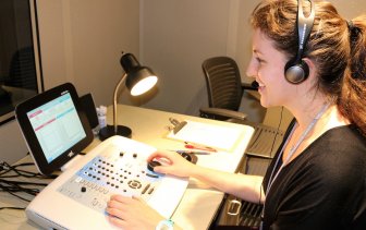 Profile of smiling graduate student with headphones on; her hands on audiometric equipment with a display screen