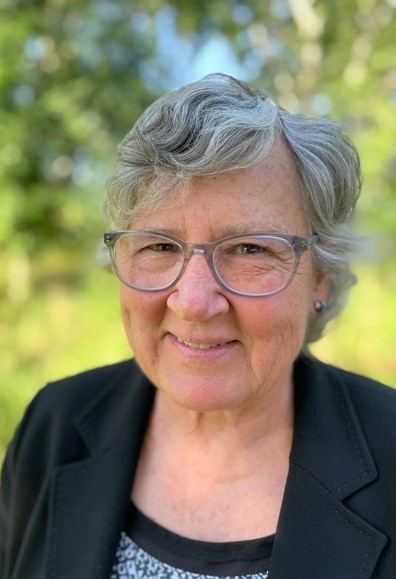 Gisele Muller-Parker smiling at the camera, with wavy gray hair, glasses, wearing a black blazer in front of a background of greenery