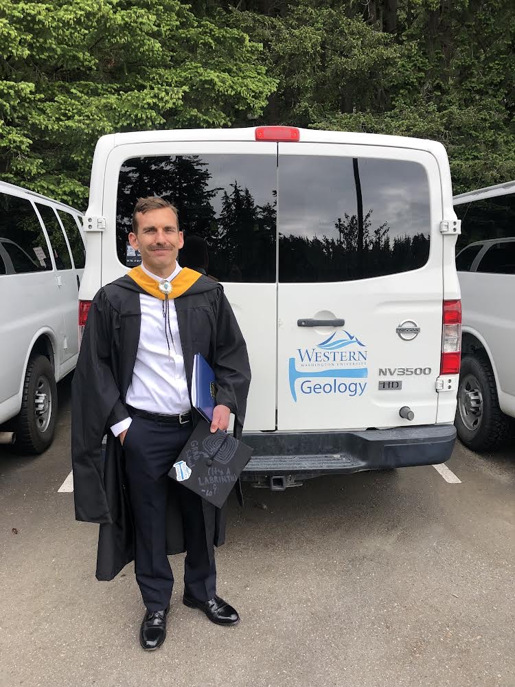 WWU graduate alum Ed Fordham standing in gown and hood next to white van with WWU logo and "Geology" on the side