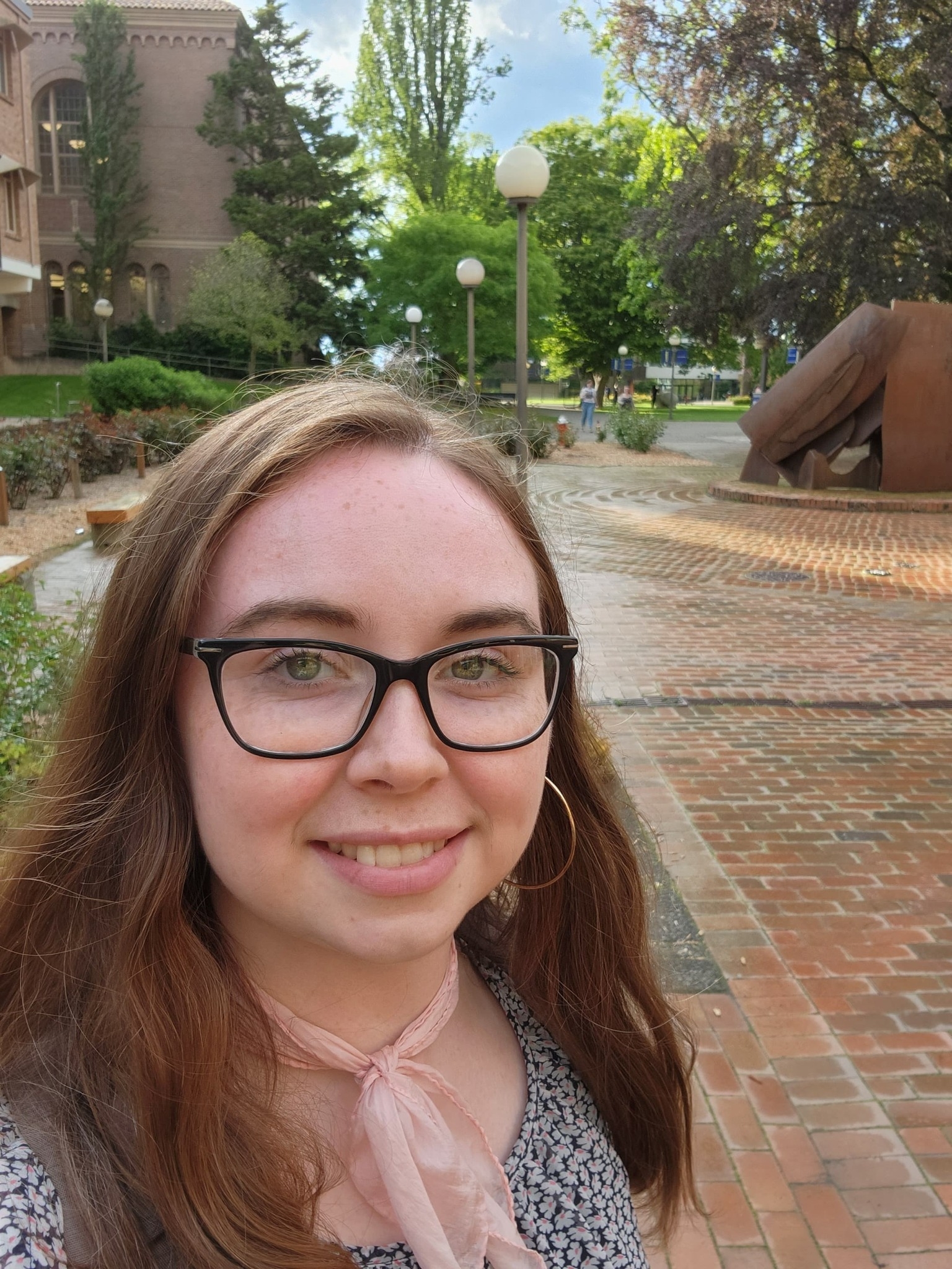 WWU outstanding graduate student Caity Scott with a brick pathway and an abstract iron sculpture in the background