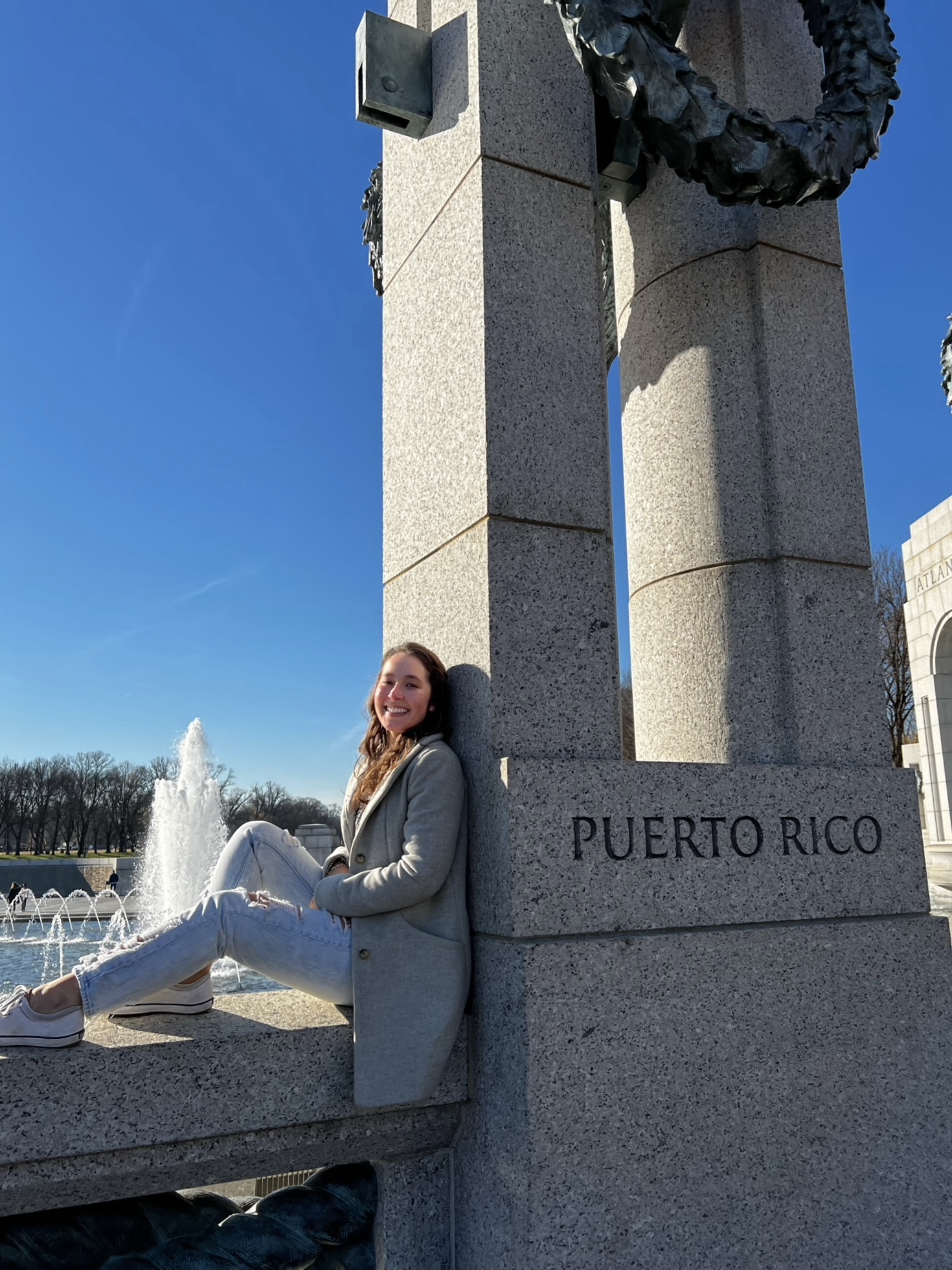 WWU outstanding graduate student Adriana Garcia-Gonzalez sitting on a ledge with legs out to left and her back up against a column that says "Puerto Rico"--fountain in background