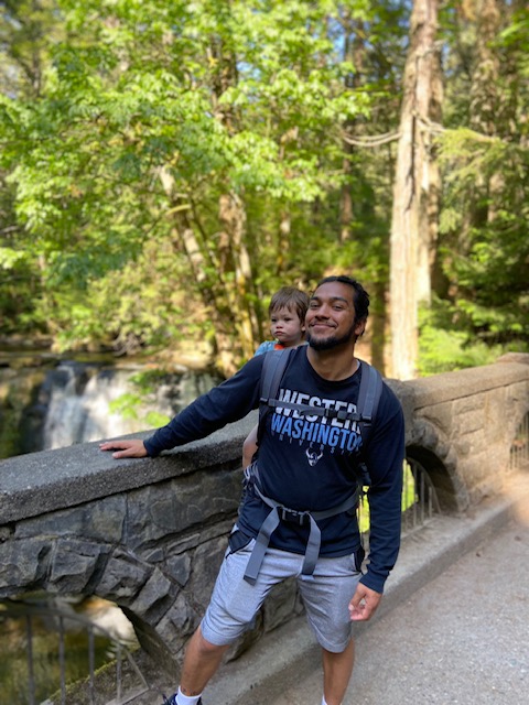 WWU Outstanding Grad Student Kosal (Junior) Gonzales with baby on his back, smiling on a bridge with waterfall and trees in the background 