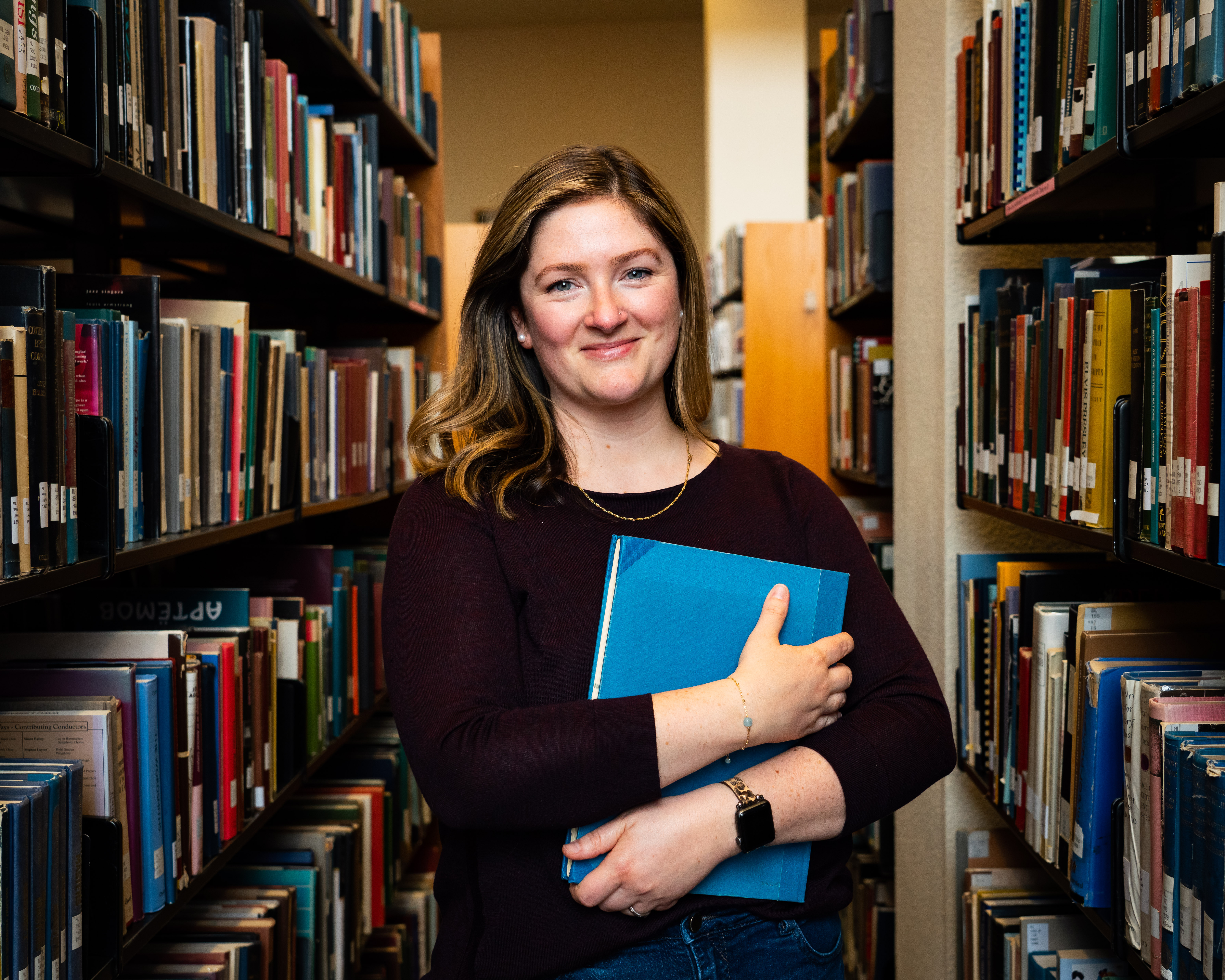 WWU graduate student of music Kyra Rengstorf holding a book to her chest with shelves of books in the background