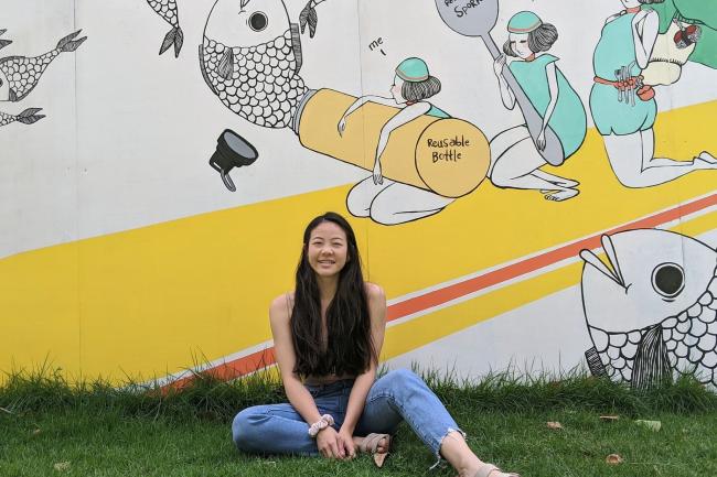 WWU graduate LiAn Noonan sitting in grass with yellow, white, and teal street art in the background