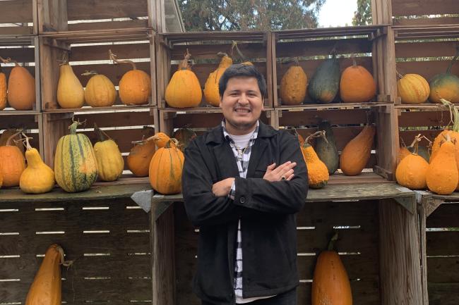 WWU graduate student Miguel Gonzalez Ramirez standing in front of shelves of pumpkins, smiling with arms crossed