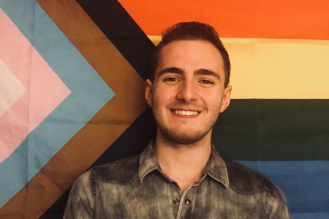 WWU graduate Andy Labay smiling with a progress pride flag in the background 