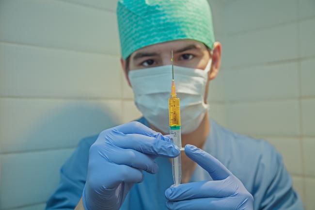 Medical provider in scrubs, gloves, cap, and mask holding a syringe out in front of their body