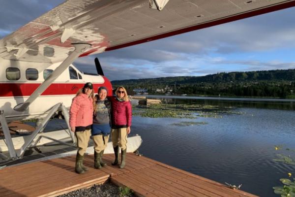 Three people bundled up in heavy coats standing on a dock prepare to board the nearby floatplane.