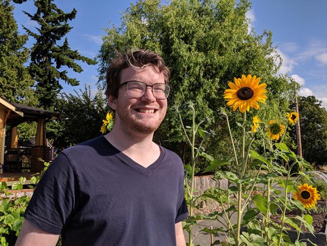 WWU graduate student Alexander Covington smiling with tall sunflowers and trees in the background