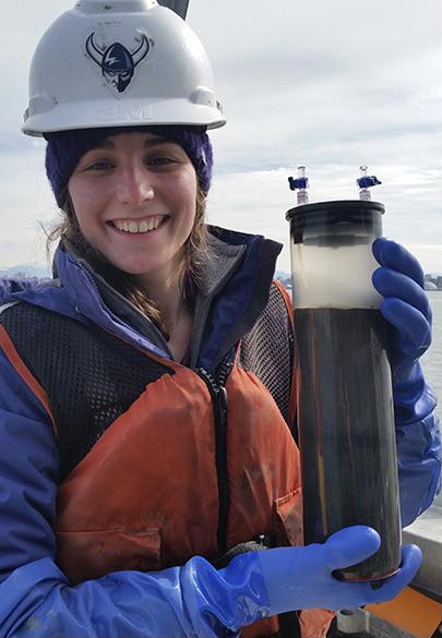 WWU graduate student Emma Rigby Santana with a hardhat on holding a large vial of dark material with clouds in the background