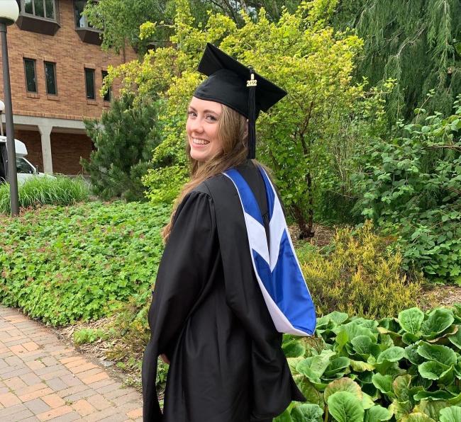 WWU outstanding graduate student Sydne Tursky in cap, gown and blue and white hood, with greenery and a brick building in the background